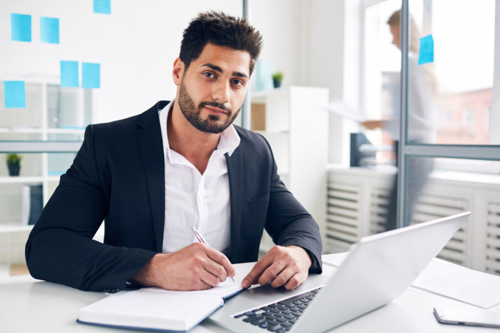 Bearded businessman wearing a suit sitting at a desk with a laptop and notepad, resting his chin on his hand and looking thoughtful