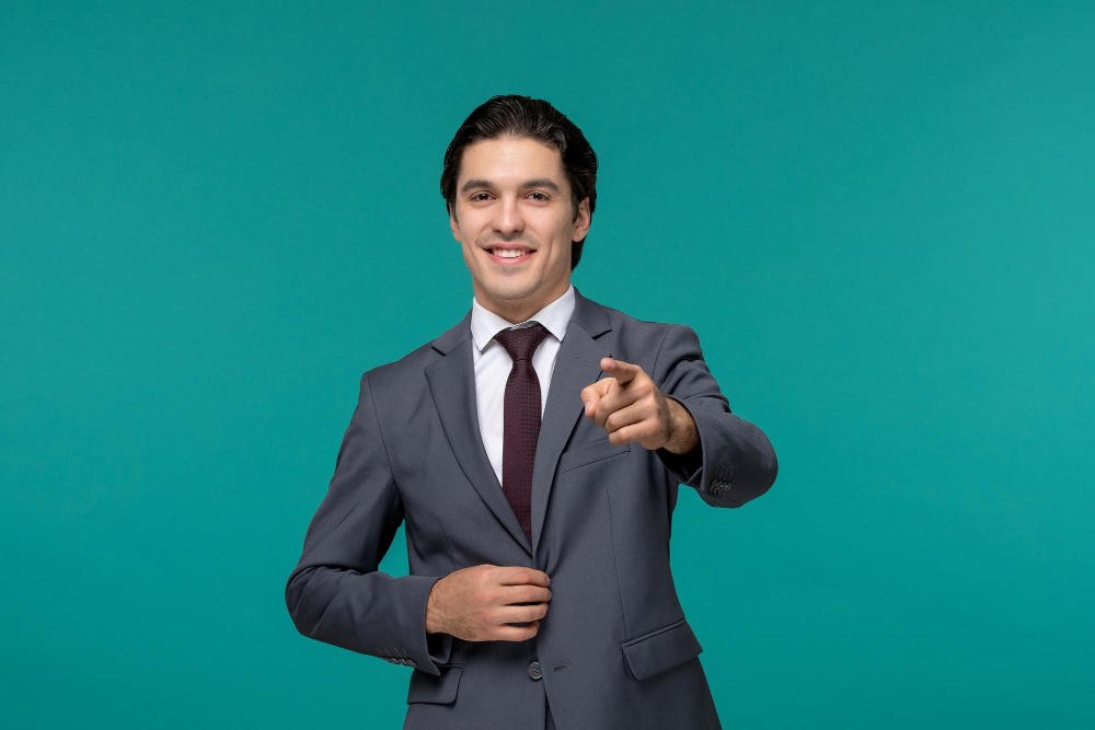 Portrait of a young man in a dark suit pointing forward and smiling against a turquoise background