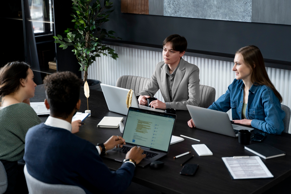 Business meeting with four colleagues around a conference table in a modern office, working on laptops and discussing a project