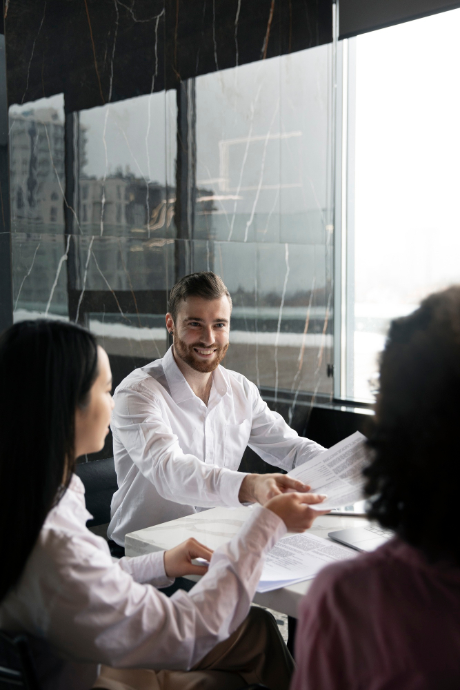 Smiling businessman in a white shirt sitting at a conference table handing a document to a female colleague, with other coworkers visible in a modern office with marble walls.