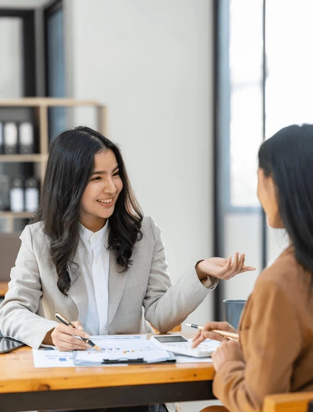 Two female colleagues in business attire discussing documents at a desk, smiling and engaged in conversation