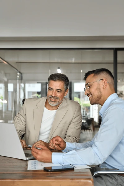 Two businessmen sitting at a table with a laptop, smiling and shaking hands to celebrate a successful deal