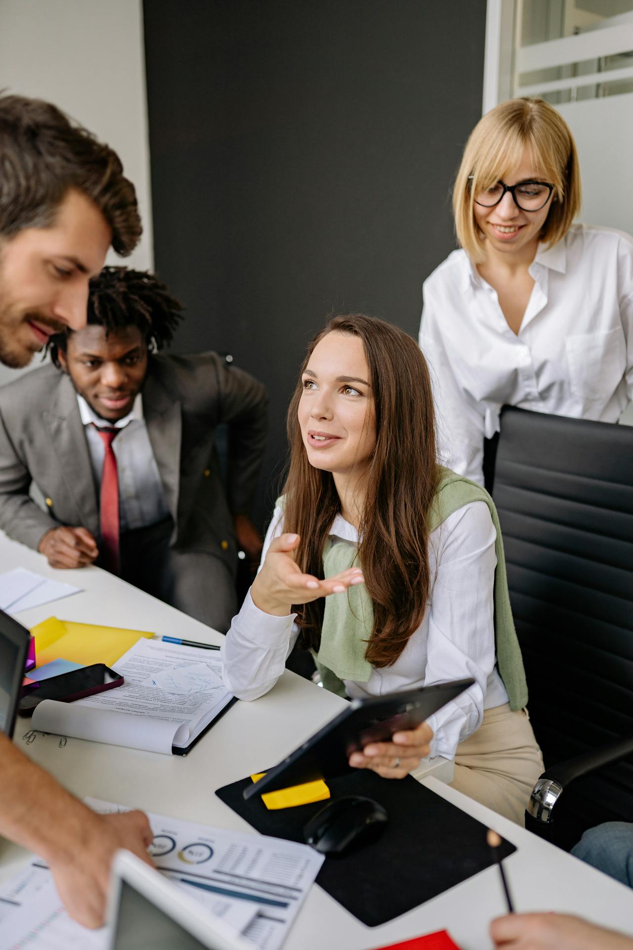 Team of business professionals collaborating at a conference table with charts and documents; a woman in a white blouse smiles while discussing ideas