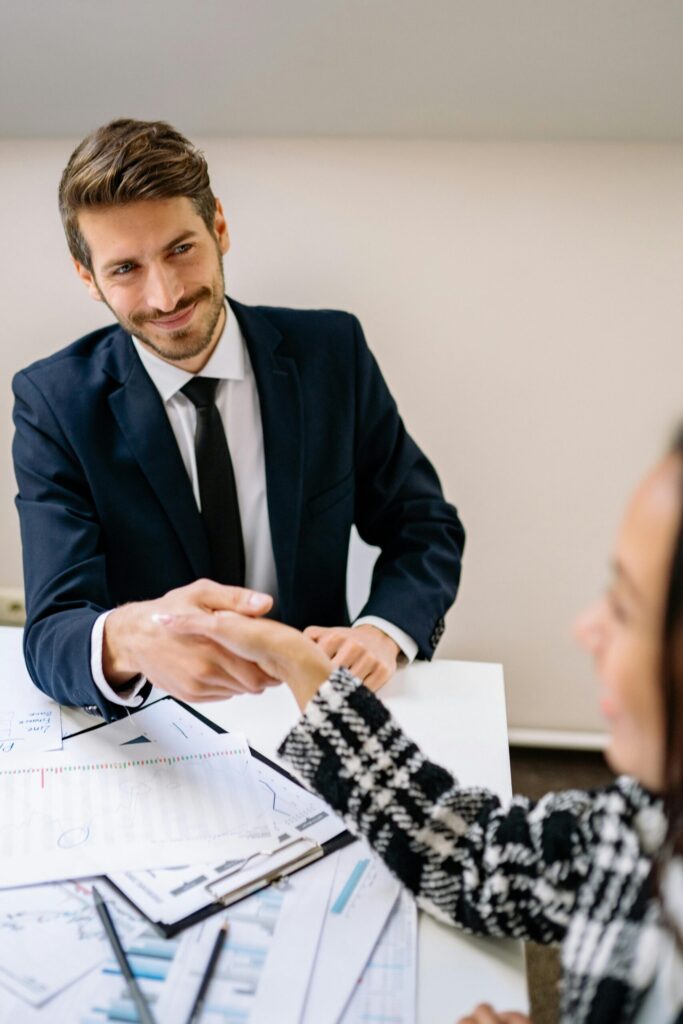 Professional businessmen shaking hands across a desk with documents and charts, smiling and wearing suits.