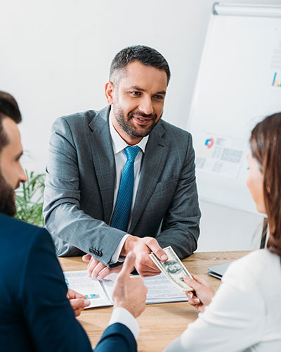 Businessman in a grey suit smiling while two colleagues hand him US dollar bills during a business meeting