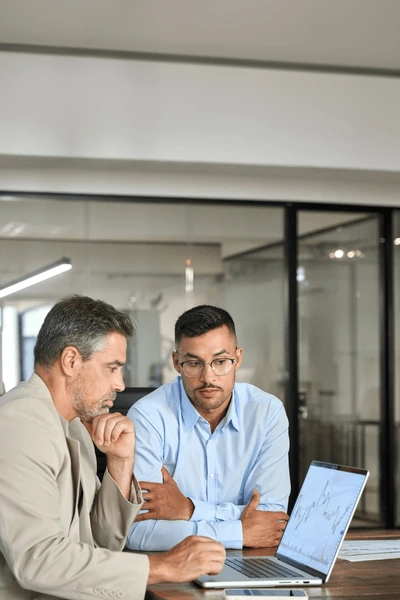 Two businessmen sitting at a table with a laptop, deep in discussion about financial documents in a modern office