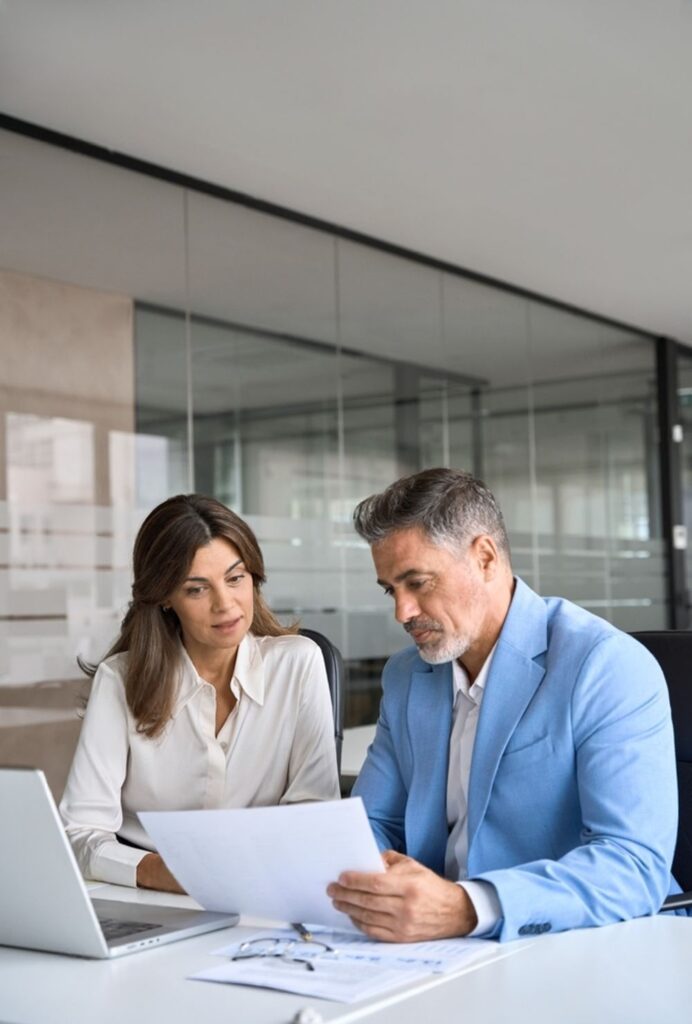 Businessman and businesswoman sitting at a desk in an office, reviewing documents together with a laptop and clipboard