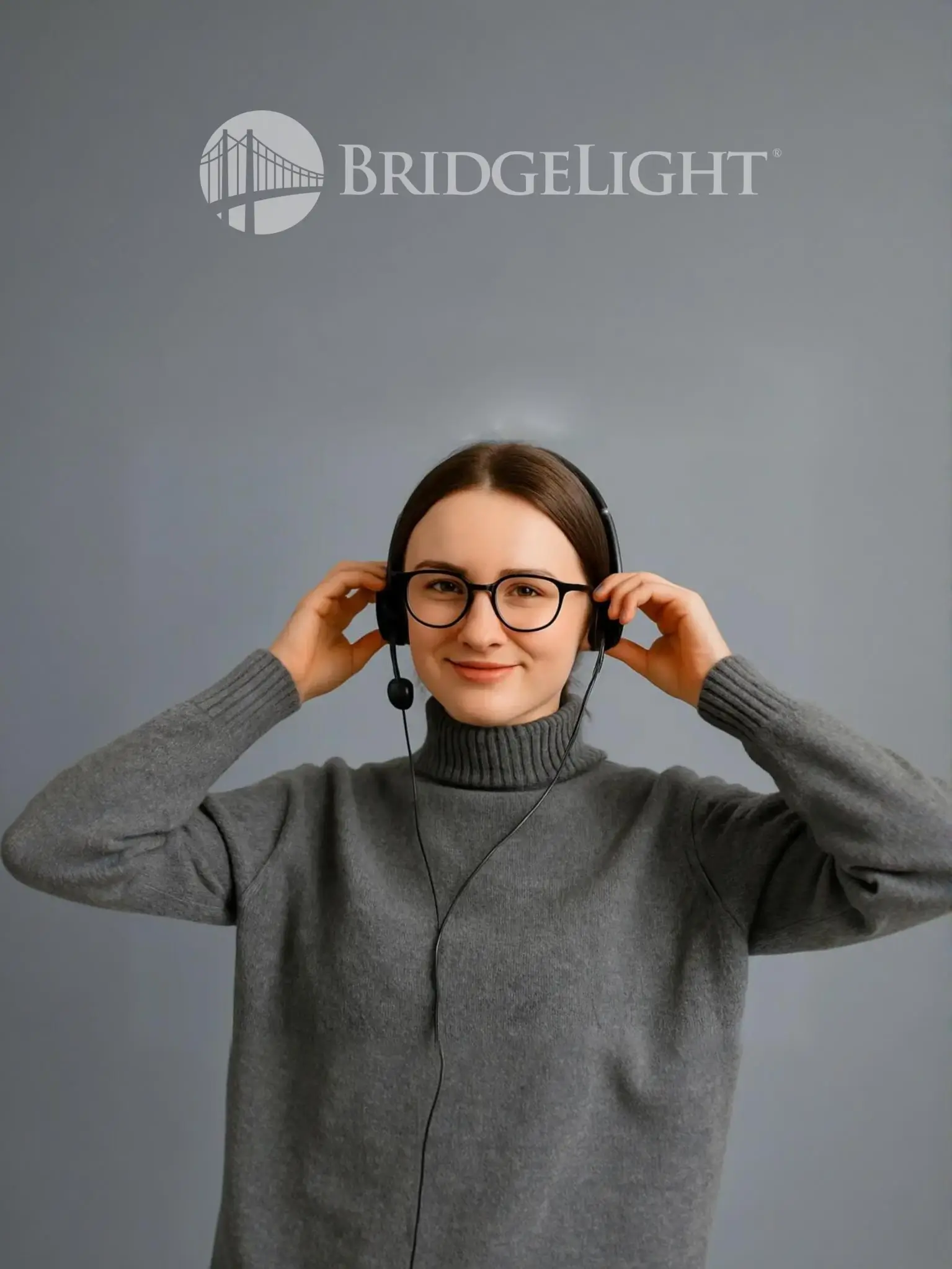 Young woman wearing glasses adjusting her eyeglasses while smiling, dressed in a grey turtleneck sweater with BridgeLight logo in the background