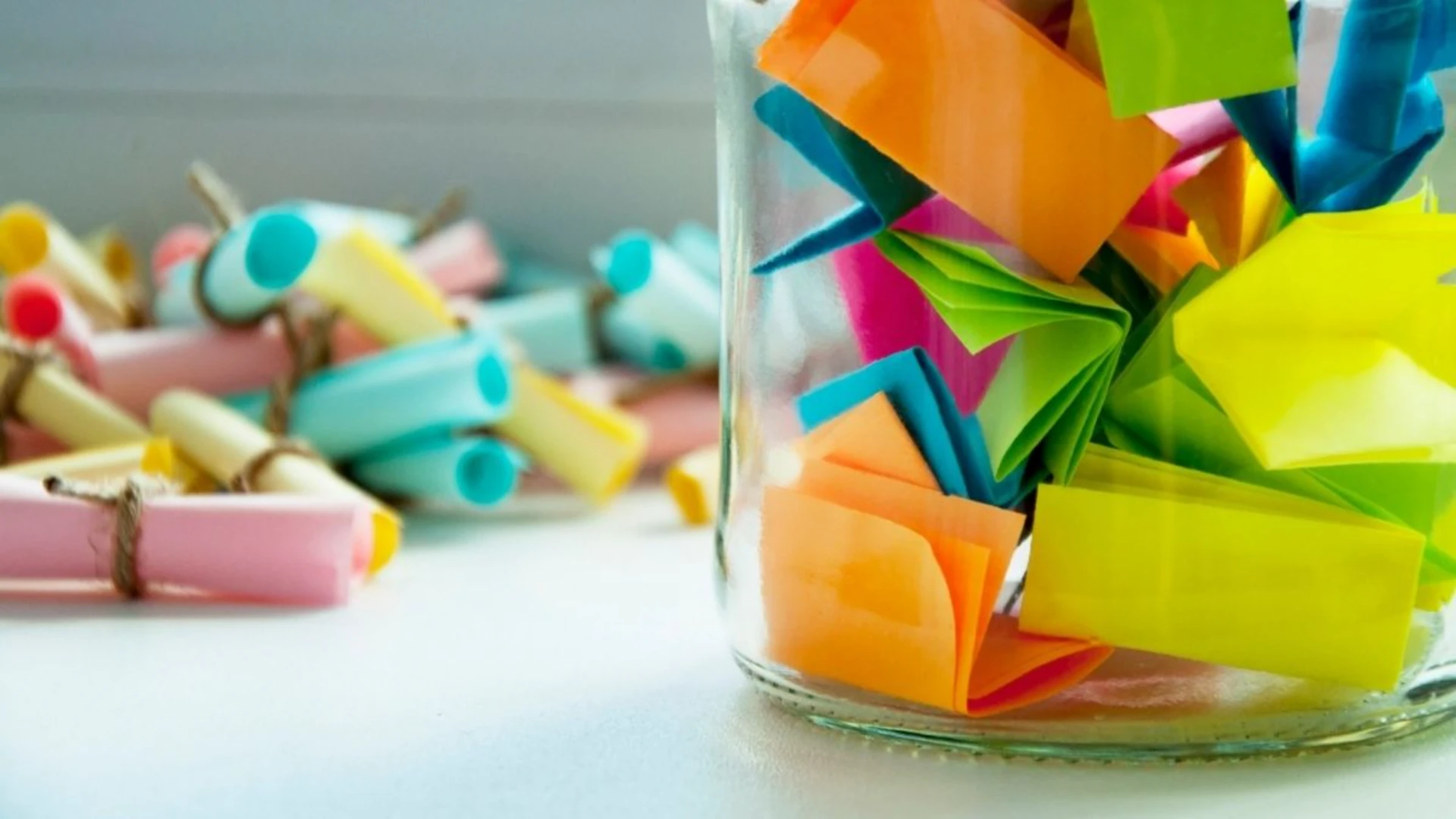glass-jar-filled-with-folded-colorful-paper-slips-with-additional-rolled-notes-tied-with-twine-in-the-background