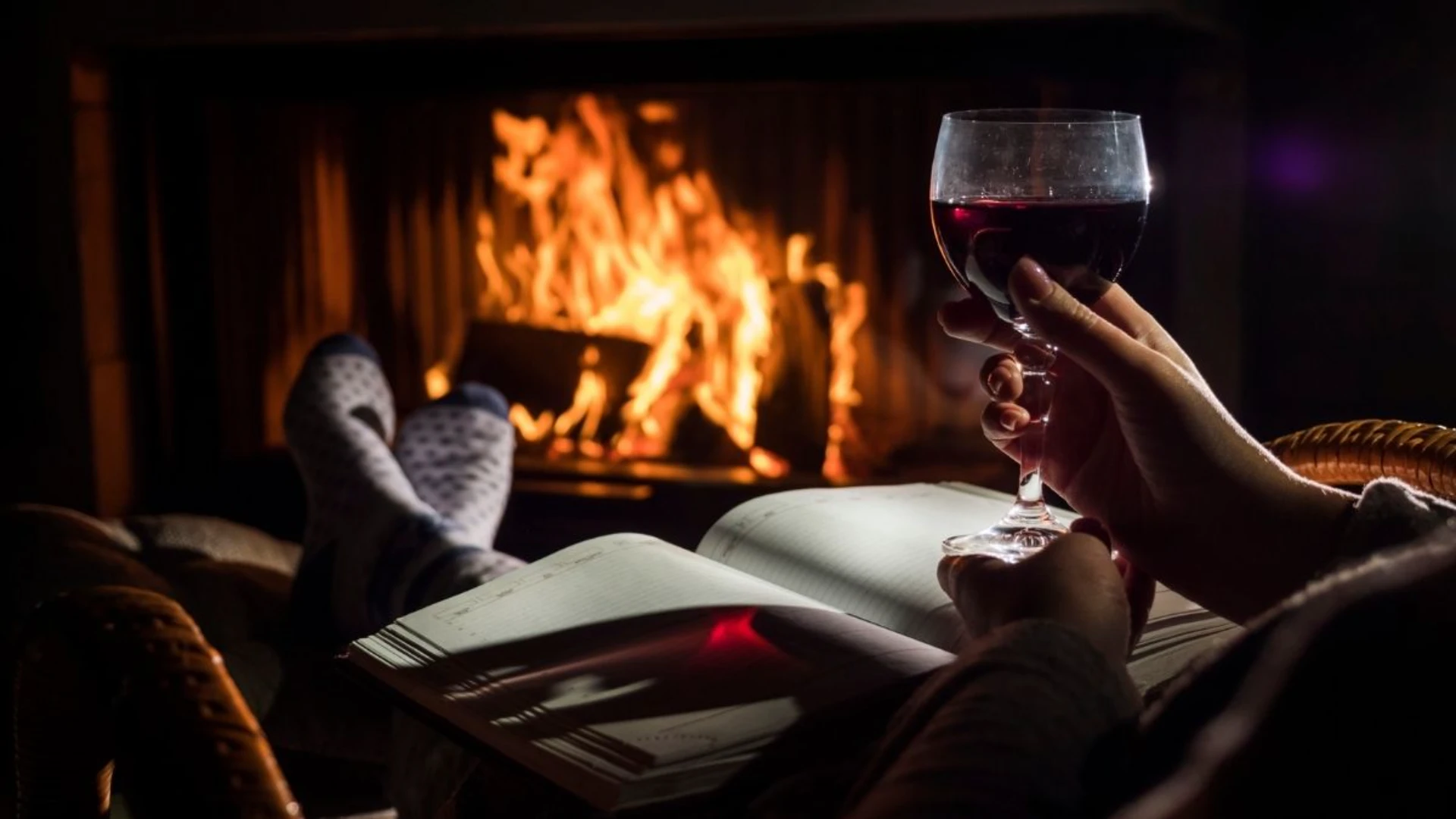 Person relaxing by a fireplace with their feet up, holding a glass of red wine and an open book